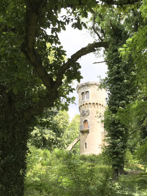 Runder Turm mit Zinnen, teilweise von grünen Bäumen und Sträuchern umgeben