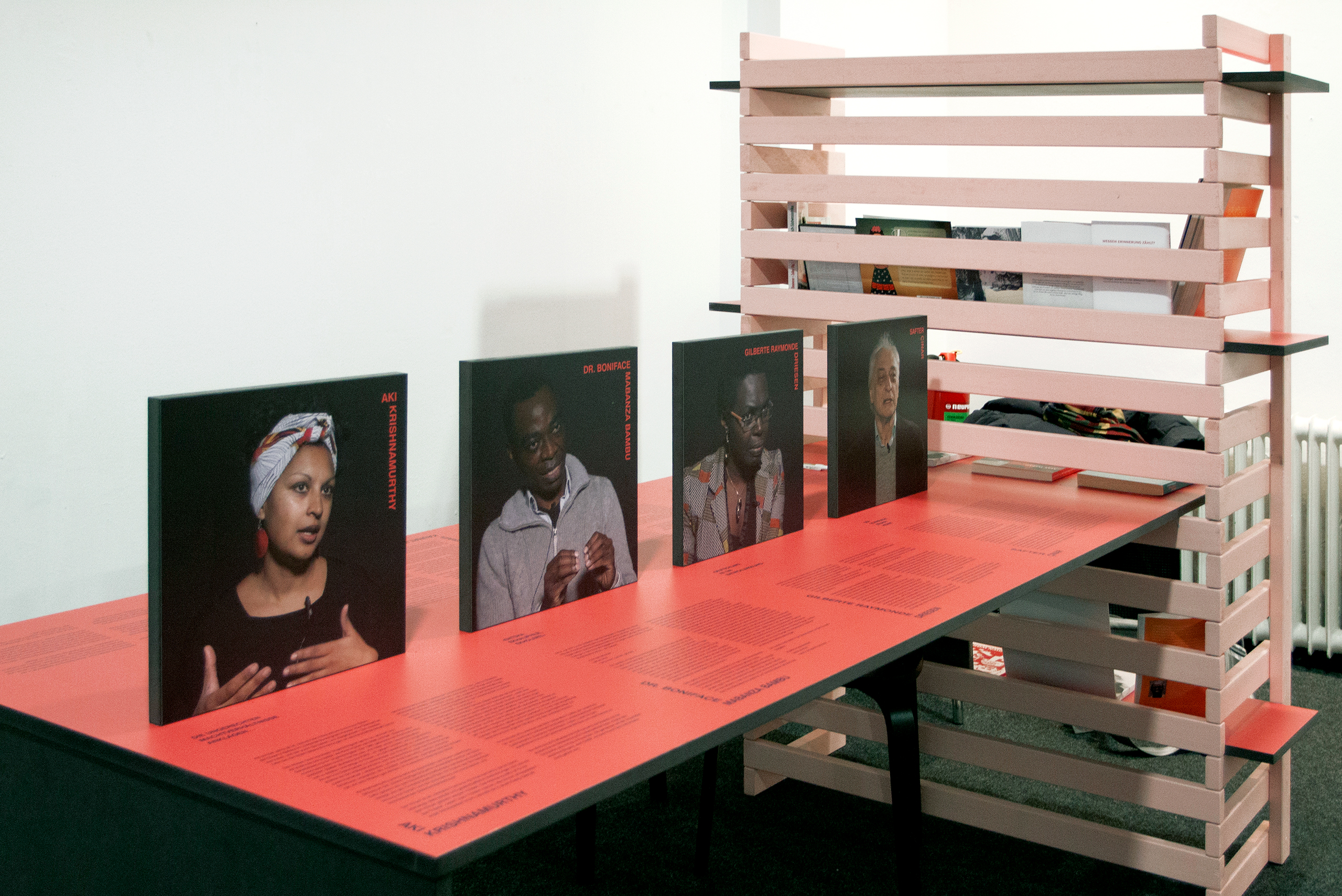 Ausstellungstisch mit vier stehenden Porträtfotos von Personen und auf dem Tisch erläuternder Text, im Hintergrund ein Holzregal mit Büchern