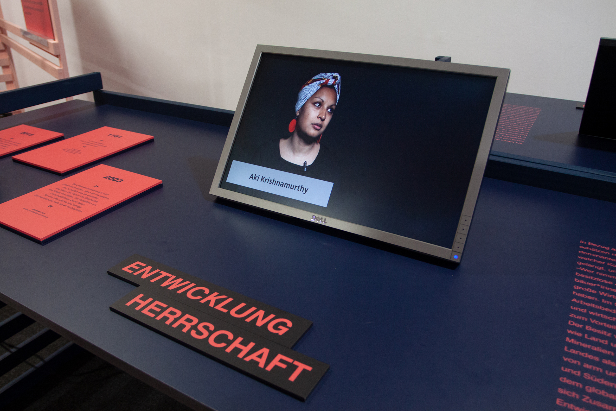 Bildschirm auf einem schwarzen Tisch mit dem Namen 'Aki Krishnamurthy' und roten Schildern mit den Worten 'ENTWICKLUNG' und 'HERRSCHAFT'