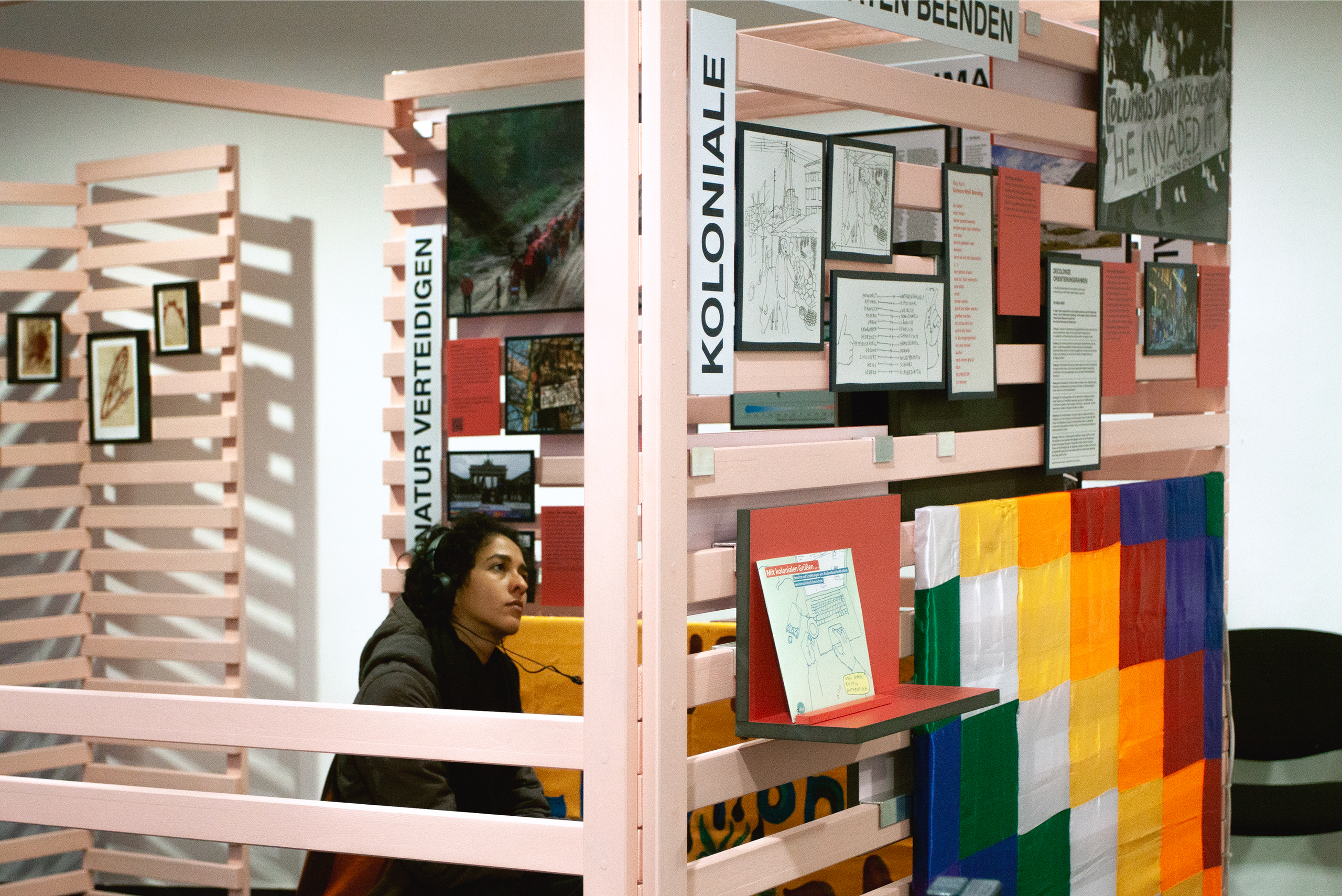 Ausstellung mit Holzlattenwänden, auf denen Bilder, Texttafeln und ein buntes Quadratmuster aus Stoff oder Papier angebracht sind. Eine Besucherin betrachtet die Ausstellung an einer Audio- oder Videostation.