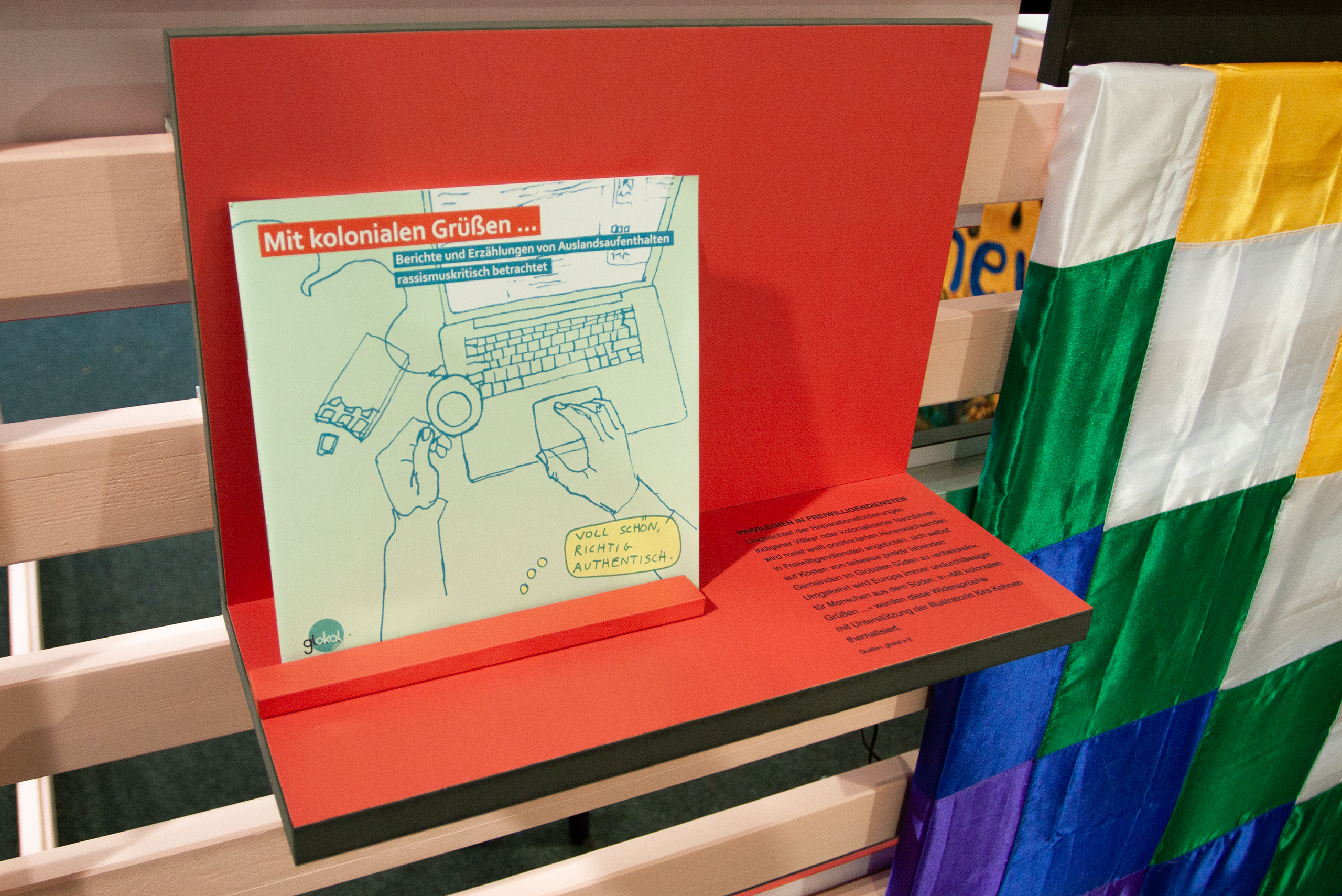 Roter Aufsteller mit einer Illustration von Händen an einer Tastatur und dem Text 'Mit kolonialen Grüßen ... Berichte und Erzählungen von Auslandsaufenthalten rassismuskritisch betrachtet'