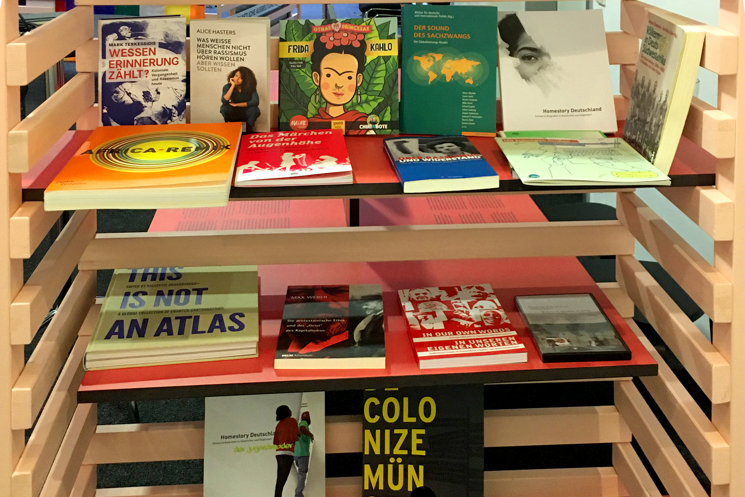 Holzregal mit mehreren Büchern, darunter Titel wie 'Wessen Erinnerung zählt', 'This is not an atlas' und 'Homestory Deutschland', einige Bücher liegen flach, andere stehen aufrecht