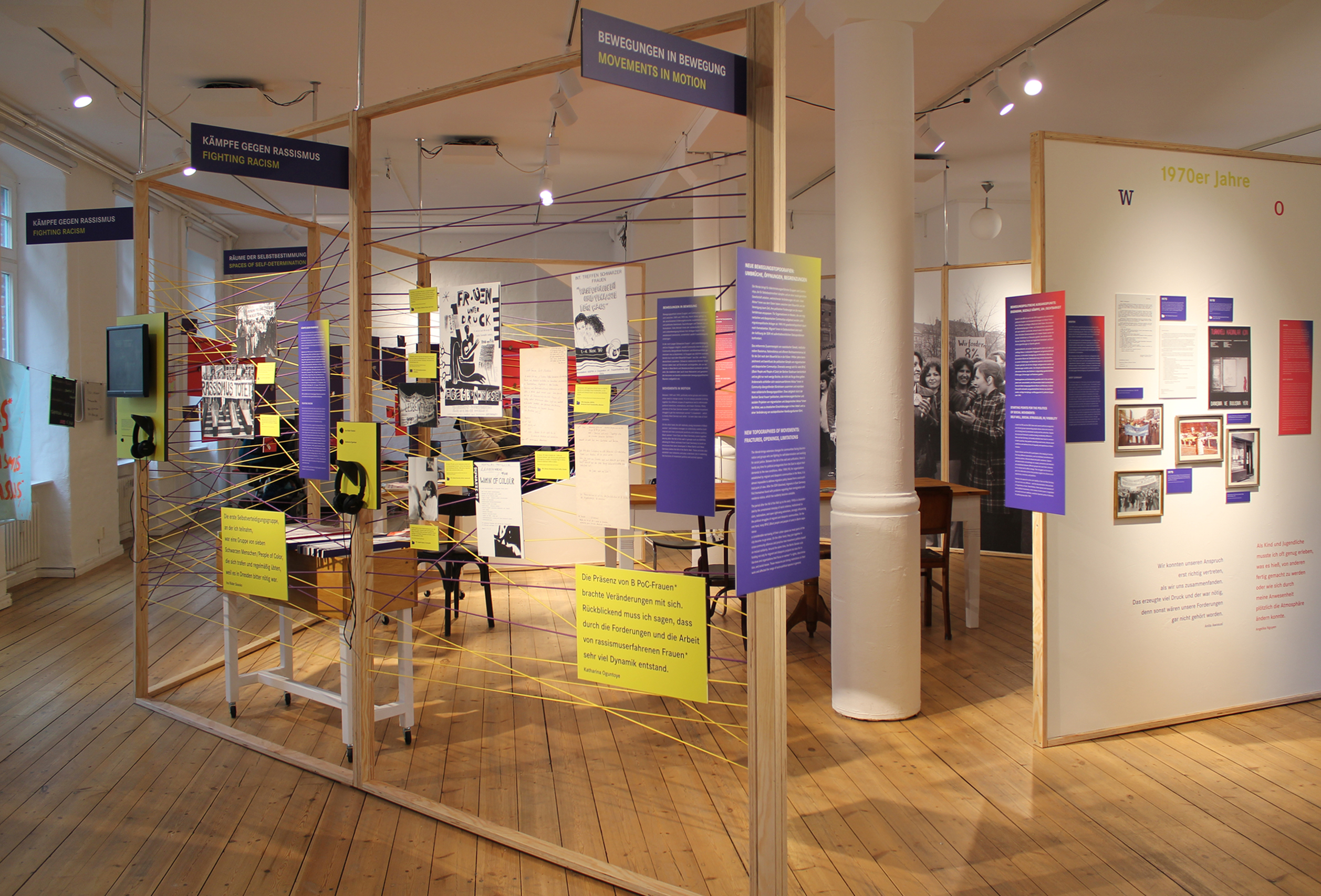 Blick in die Ausstellung mit mehreren gelben und violetten Infotafeln, die an einem Holzrahmen mit ebenfalls gelben und violetten Fäden befestigt sind.