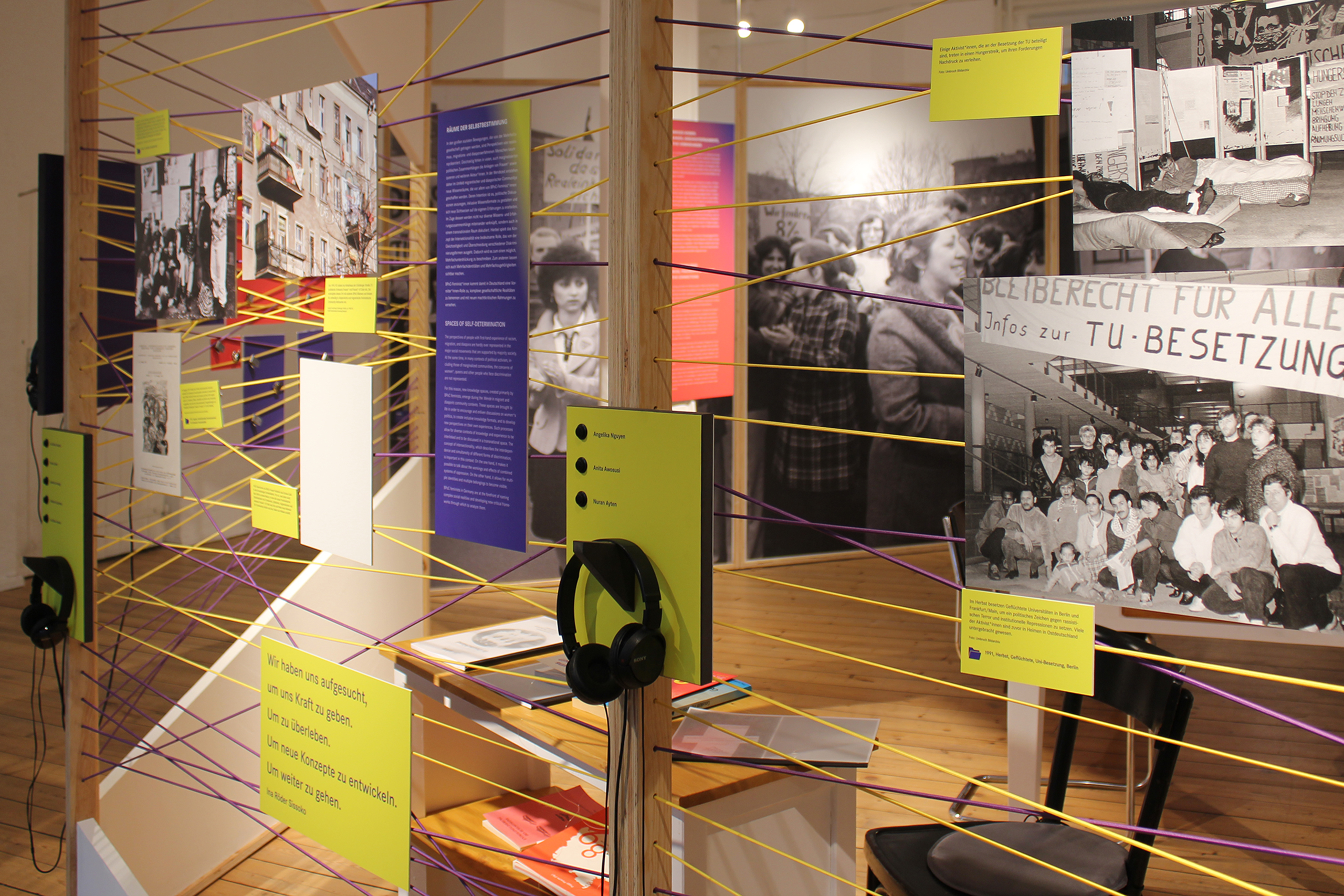 Ausstellung mit schwarz-weißen Fotografien, Texttafeln und Hörstationen mit Kopfhörern, verbunden durch gelbe und violette Fäden.