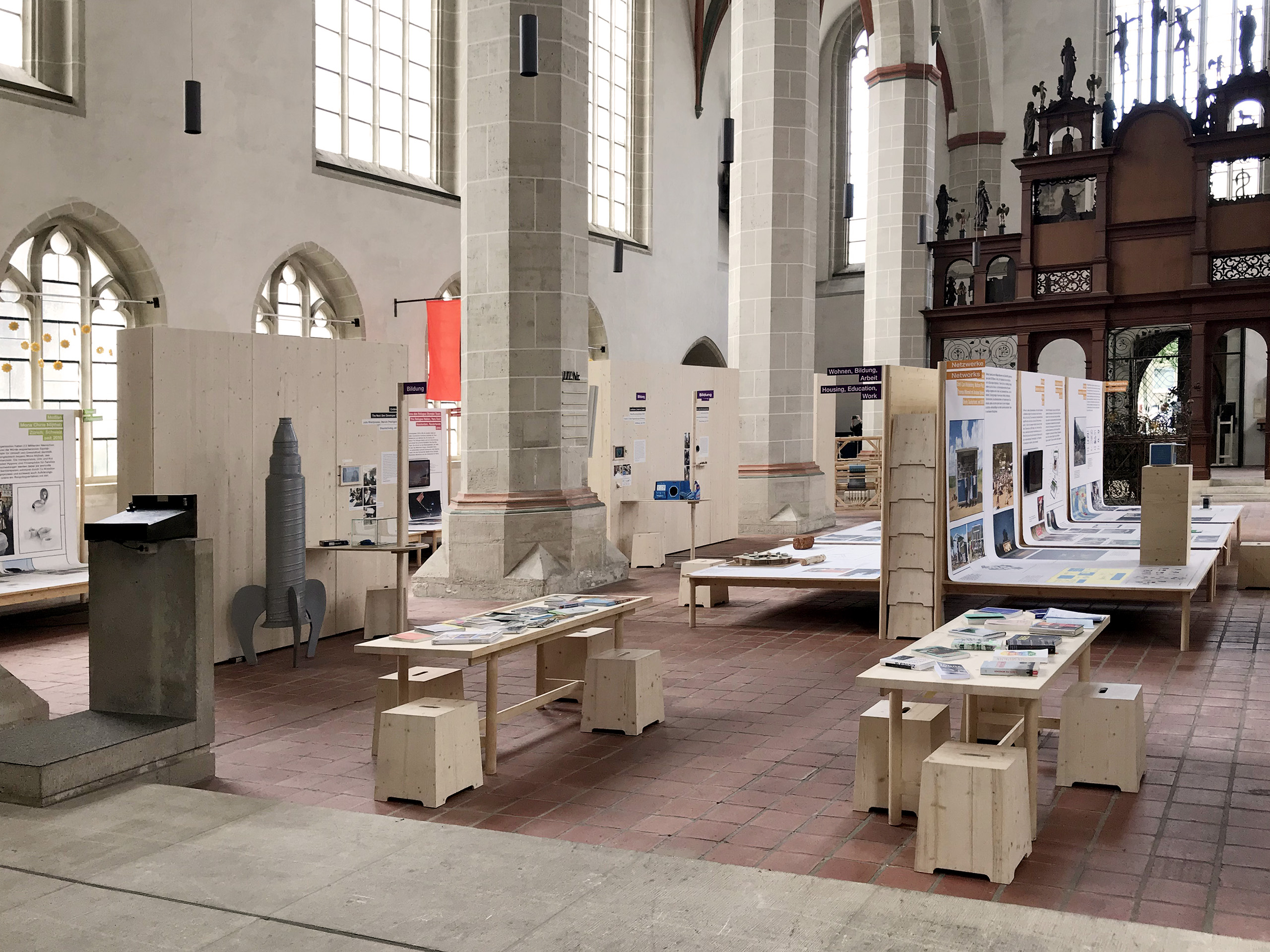 Blick in die Ausstellung im Innenraum einer Kirche mit zahlreichen Ausstellungsmodulen aus Holz wie Ausstellungstafeln, Tischen mit Hockern und Wegweisern