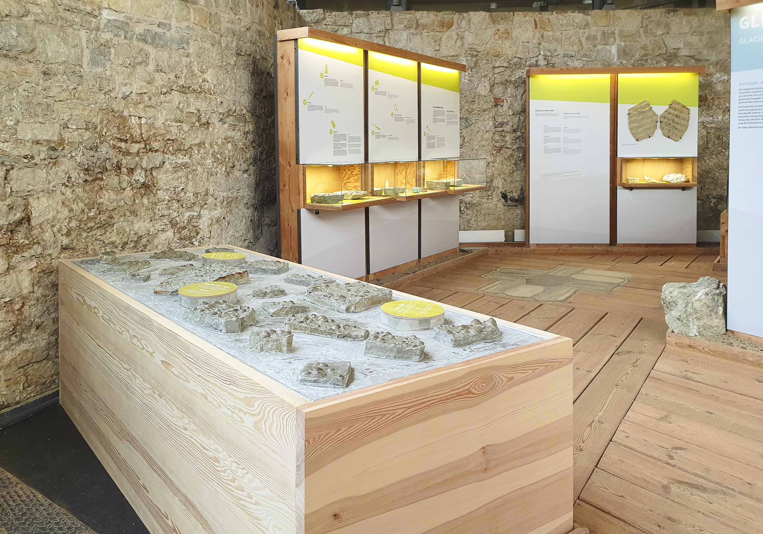 Blick in die Ausstellung mit Fossilientisch, beleuchteten Vitrinen und Informationstafeln in einem Raum mit Steinmauern und Holzboden.