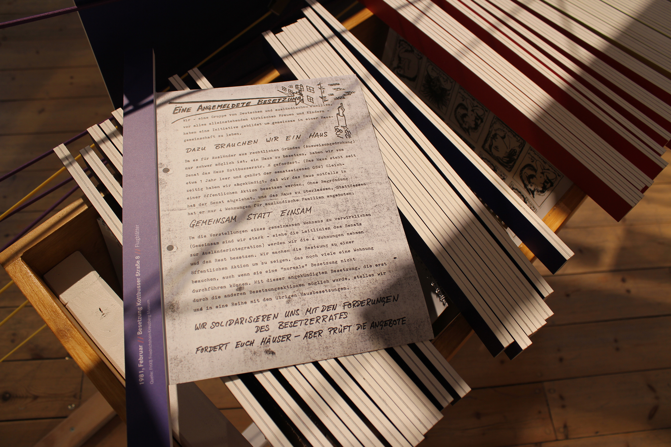 Zettel mit Text liegt auf einem Holzgestell mit vielen schmalen Holzlatten, Sonnenlicht fällt schräg darauf.