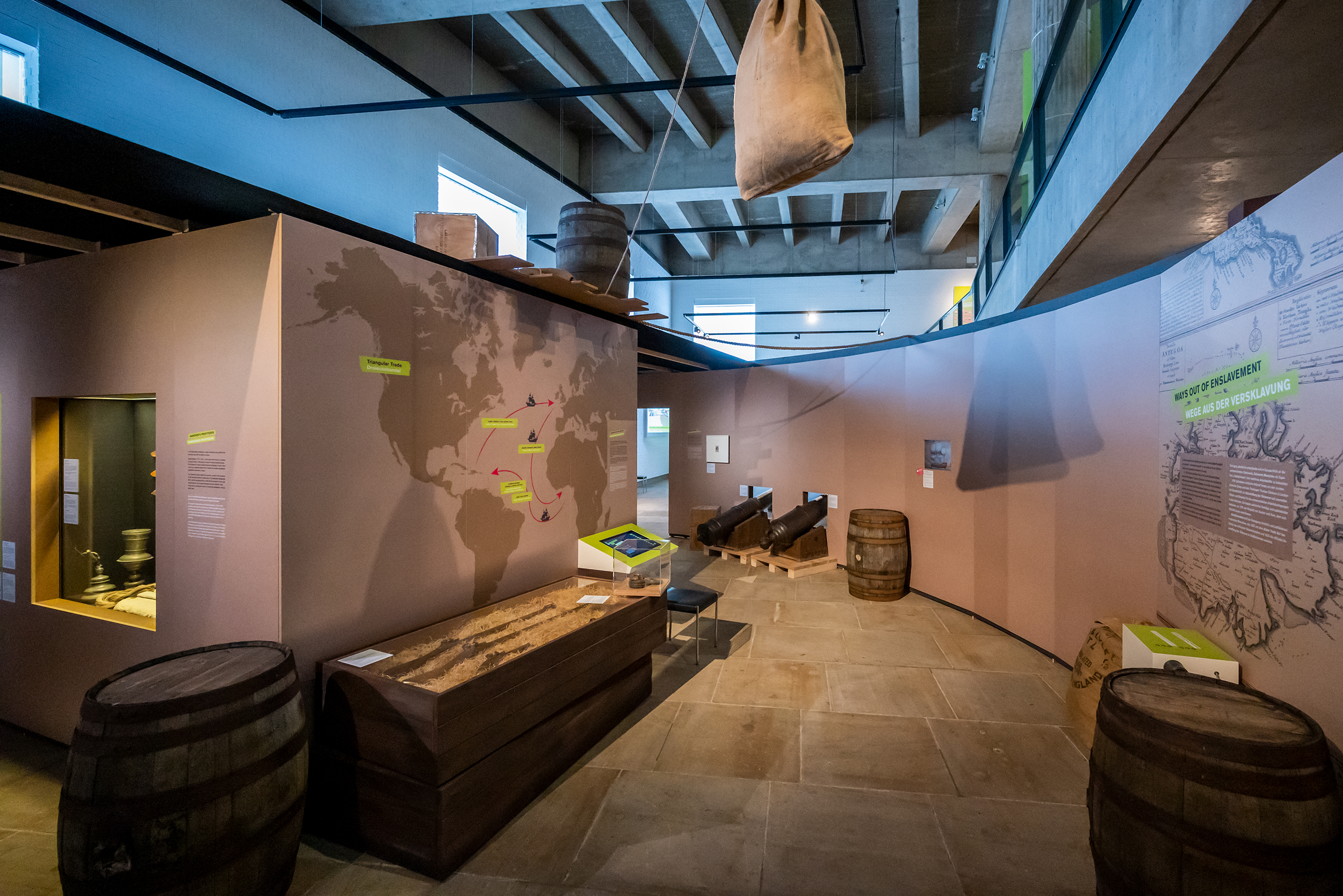 Blick in die Ausstellung, links eine Weltkarte zum Thema "Dreieckshandel" an der Wand. Holzfässer, eine Holzkiste mit Gewehren, zwei Kanonen und ein Leinensack schaffen die Atmosphäre eines Handelsschiffs aus dem 18. Jahrhundert.