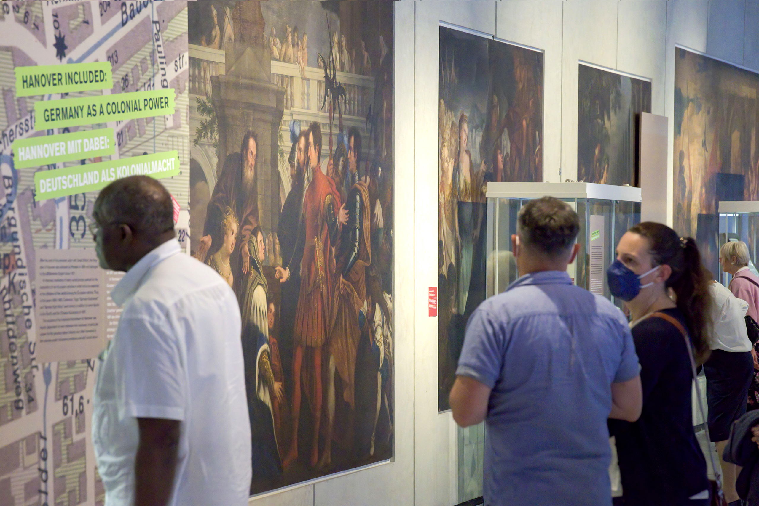 Mehrere Besucher:innen betrachten Ausstellungswände mit Gemälden und Texten zu "Deutschland als Kolonialmacht"