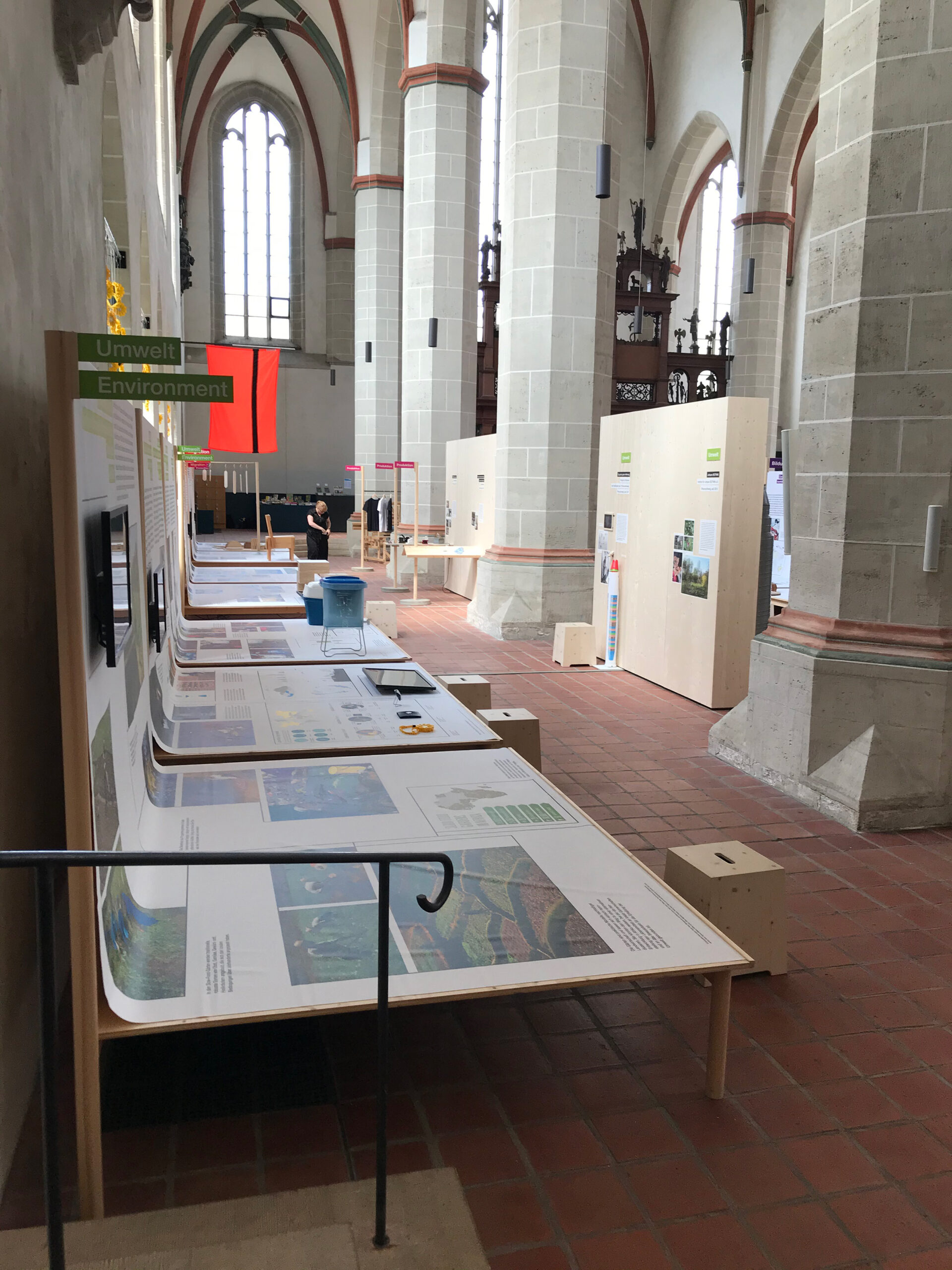 Ausstellung mit Infotafeln, zum Beispiel zu den Themen Umwelt und Produktion, in einem hohen, gotischen Kirchenraum mit Säulen und Spitzbogenfenstern.