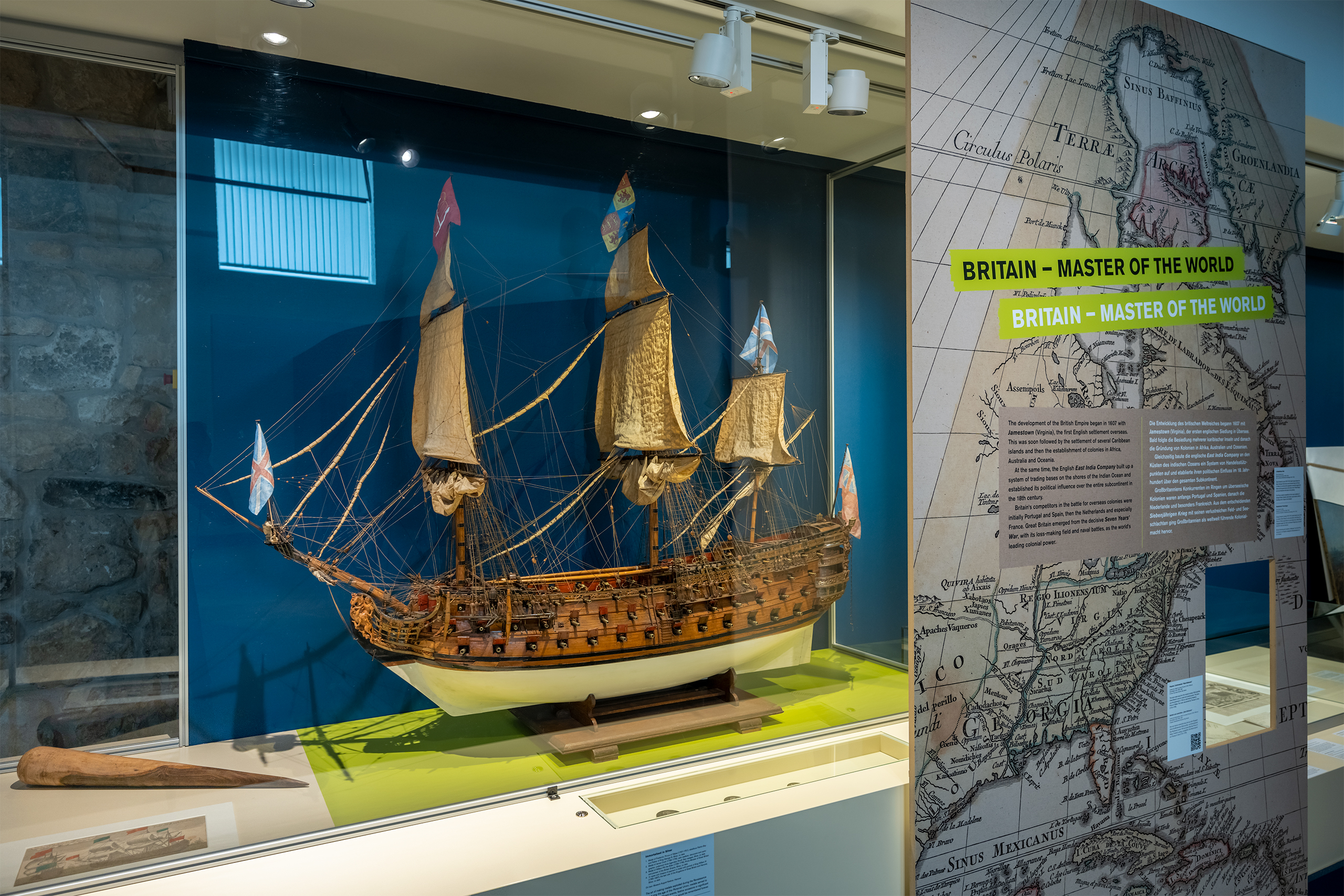 Modell eines historischen Segelschiffs mit drei Masten in einer Vitrine neben einer Wand mit einer Landkarte und dem Text 'BRITAIN - MASTER OF THE WORLD'.