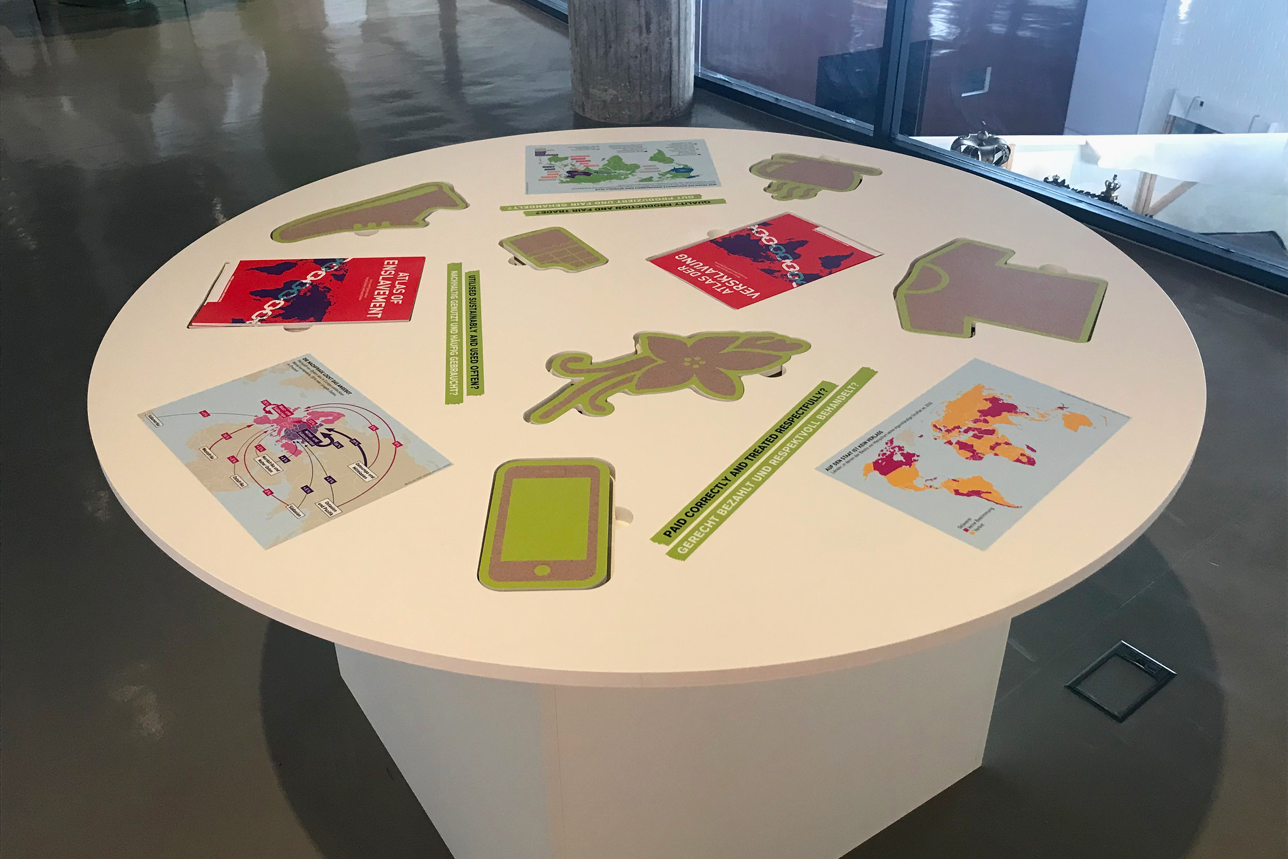 Runder weißer Tisch mit mehreren Landkarten und Fragen rund um das Thema Handel. Ein "Atlas der Versklavung" sowie verschiedene Handelsobjekte (Kleidung, Genussmittel, Elektronik) sind als herausnehmbare Objekte aus Holz in den Tisch eingelassen.