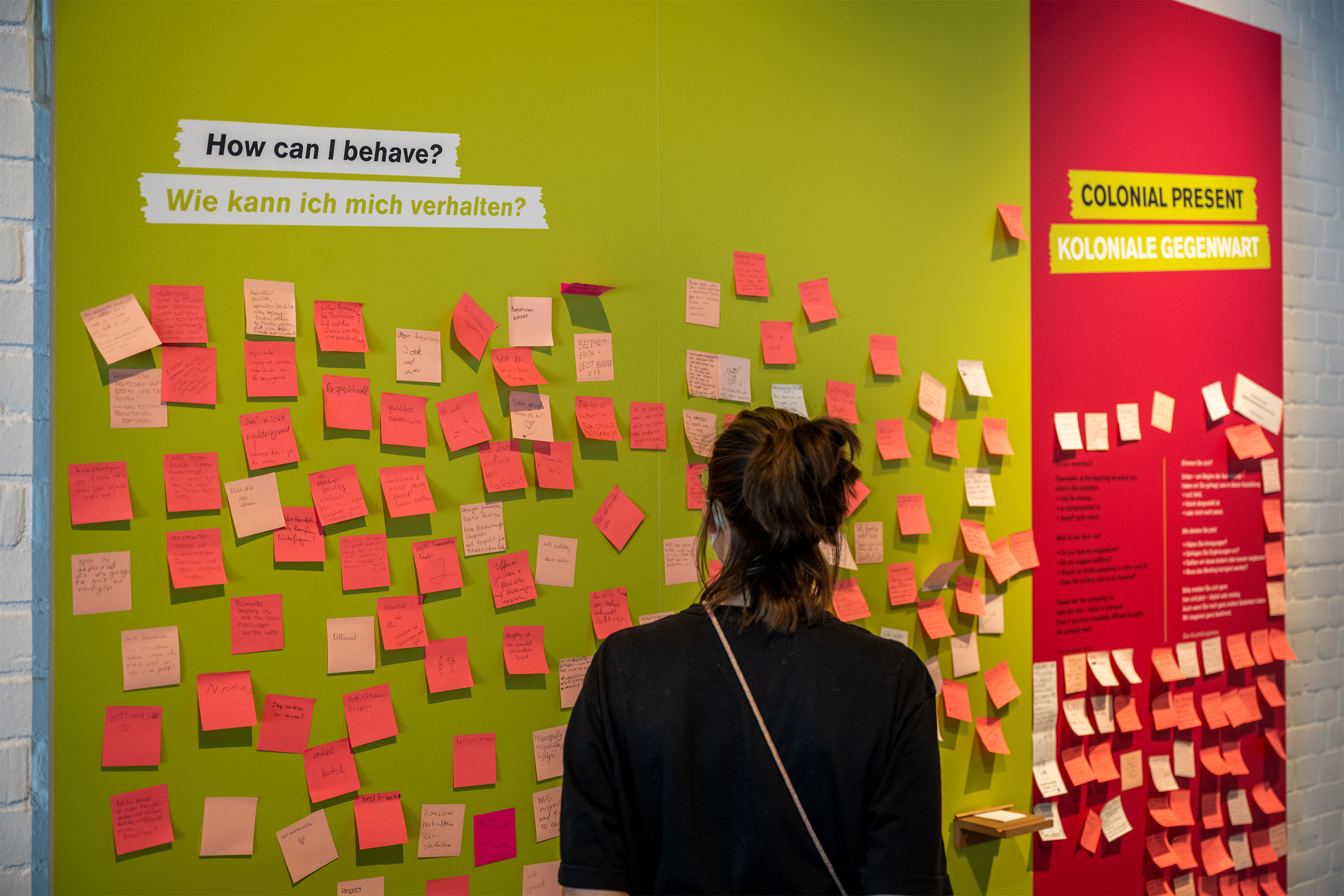 Eine Person steht vor einer Wand mit der Frage 'Wie kann ich mich verhalten?' Besucher:innen haben rote Notizzettel darunter geklebt, mit Antworten wie beispielsweise "Respektvoll", "Konsumverhalten einschränken" oder "Sei doch einfach kein Arschloch"