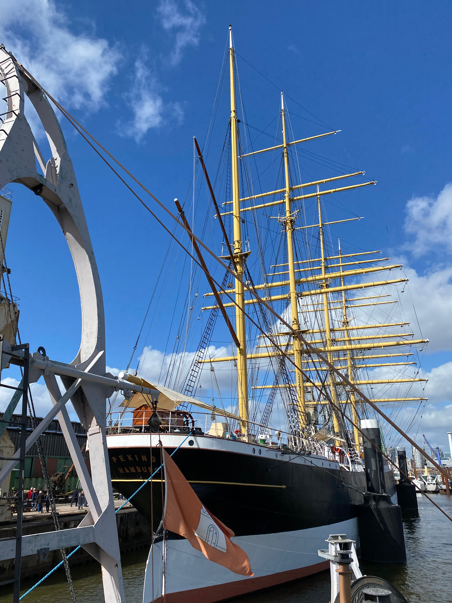 Segelschiff mit mehreren gelben Masten und weißen Segeln am Kai unter blauem Himmel