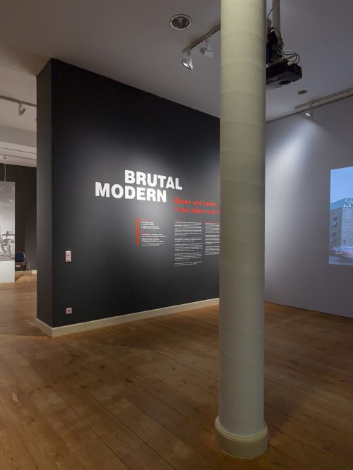 Blick in die Ausstellung. Der Titel "Brutal Modern" in weißer Schrift an schwarzer Wand im Raum, ein großflächiges Foto eines Gebäudes an der Nebenwand. In der Mitte des Raumes eine große graue Säule, im Hintergrund Blick in einen anderen Bereich mit dem Titel "Hoffnung auf die Zukunft"