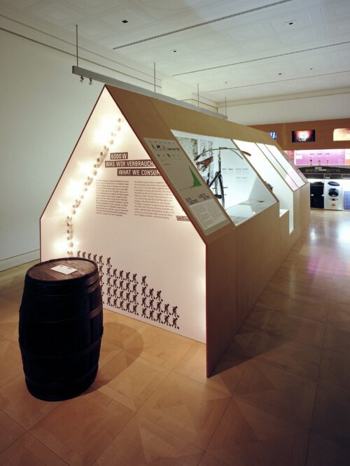 Ausstellungsraum mit beleuchtetem, hausförmigem Infostand über Energieverbrauch und einem Fass im Vordergrund