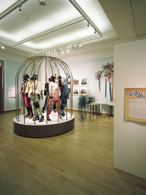 Ausstellung mit mehreren bunt gekleideten Schaufensterpuppen in einem runden, goldfarbenen Käfig auf einem Podest in einem hellen Raum mit Holzboden und Texttafeln an den Wänden