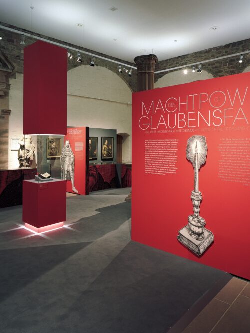 Roter Ausstellungsbereich mit großem Textpaneel 'Macht das Glaubens Power of Faith' und Abbildung einer Kerze im Ständer auf einem Buch in einem historischen Museumssaal mit Skulpturen und Gemälden.