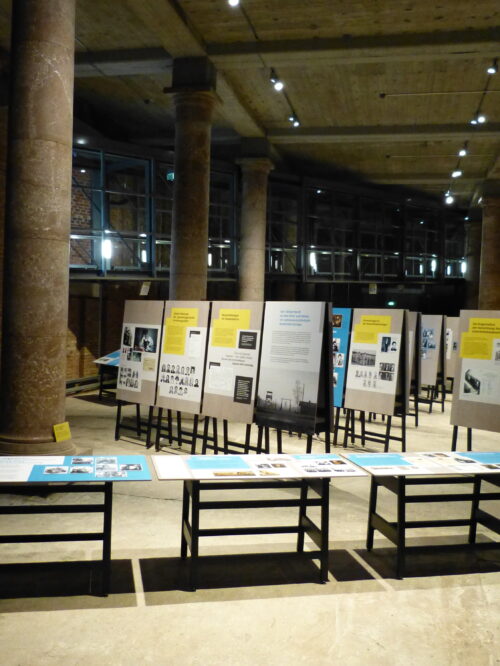 Blick in die Ausstellung mit Informationstafeln und Bildern in einem großen Raum mit Säulen