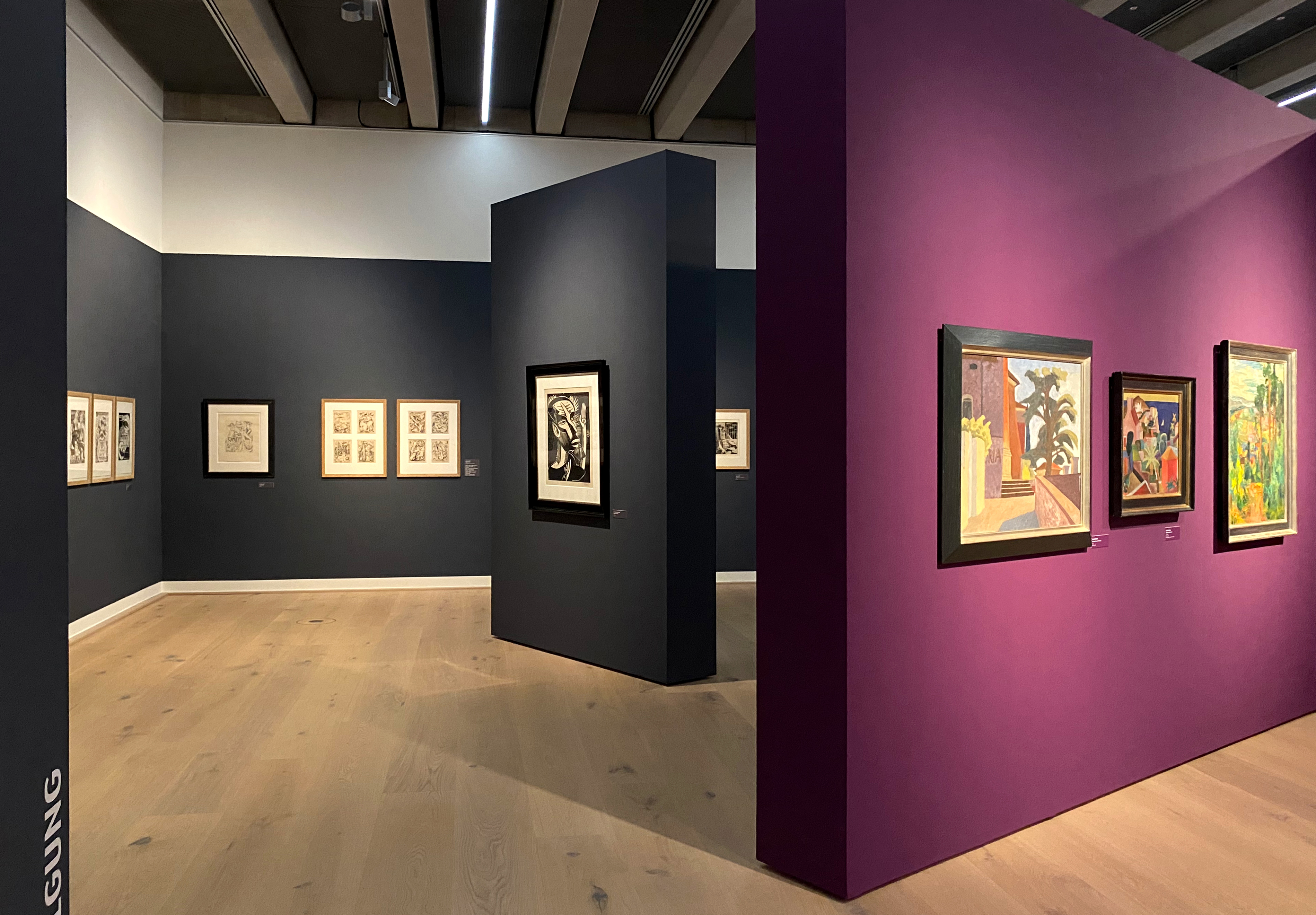 Lila und dunkelgrau-blaue Wände mit verschieden großen und farbigen Bildern. Während auf der in den Blick ragenden Wand bunte Motive zu sehen sind, sind auf den dunkelgraublauen Wänden vorrangig unbunte Werke zu sehen.