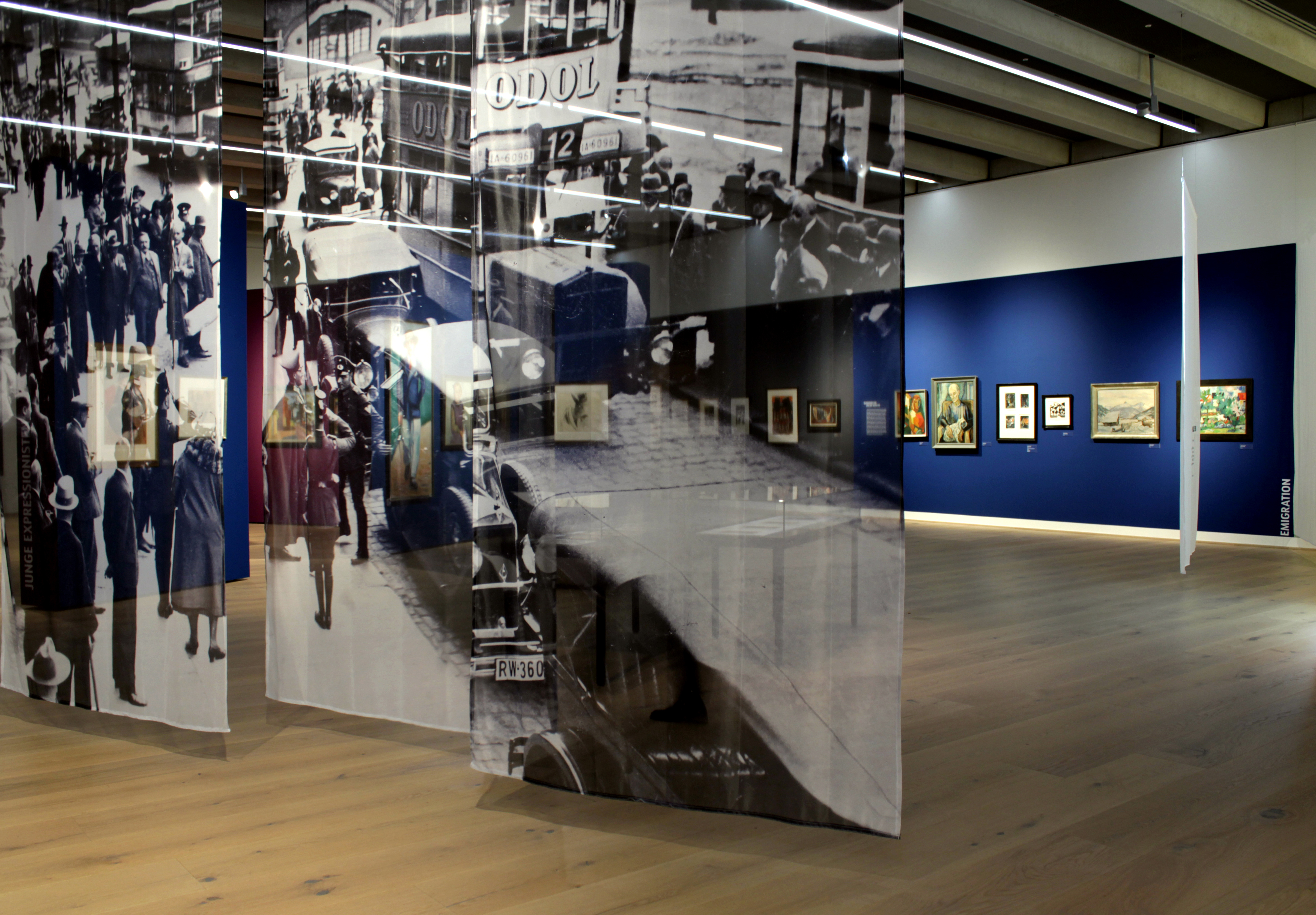 Drei hohe, schmale Transparente hängen versetzt von der Decke. Das über alle drei Gazen bedruckte Motiv ist ein schwarz-weiß Foto. Es zeigt eine alte Straßenszene voller Menschen, Autos und Busse. 'EMIGRATION' steht gestürzt in Weiß auf einer blauen Wand mit Bildern im Hintergrund.