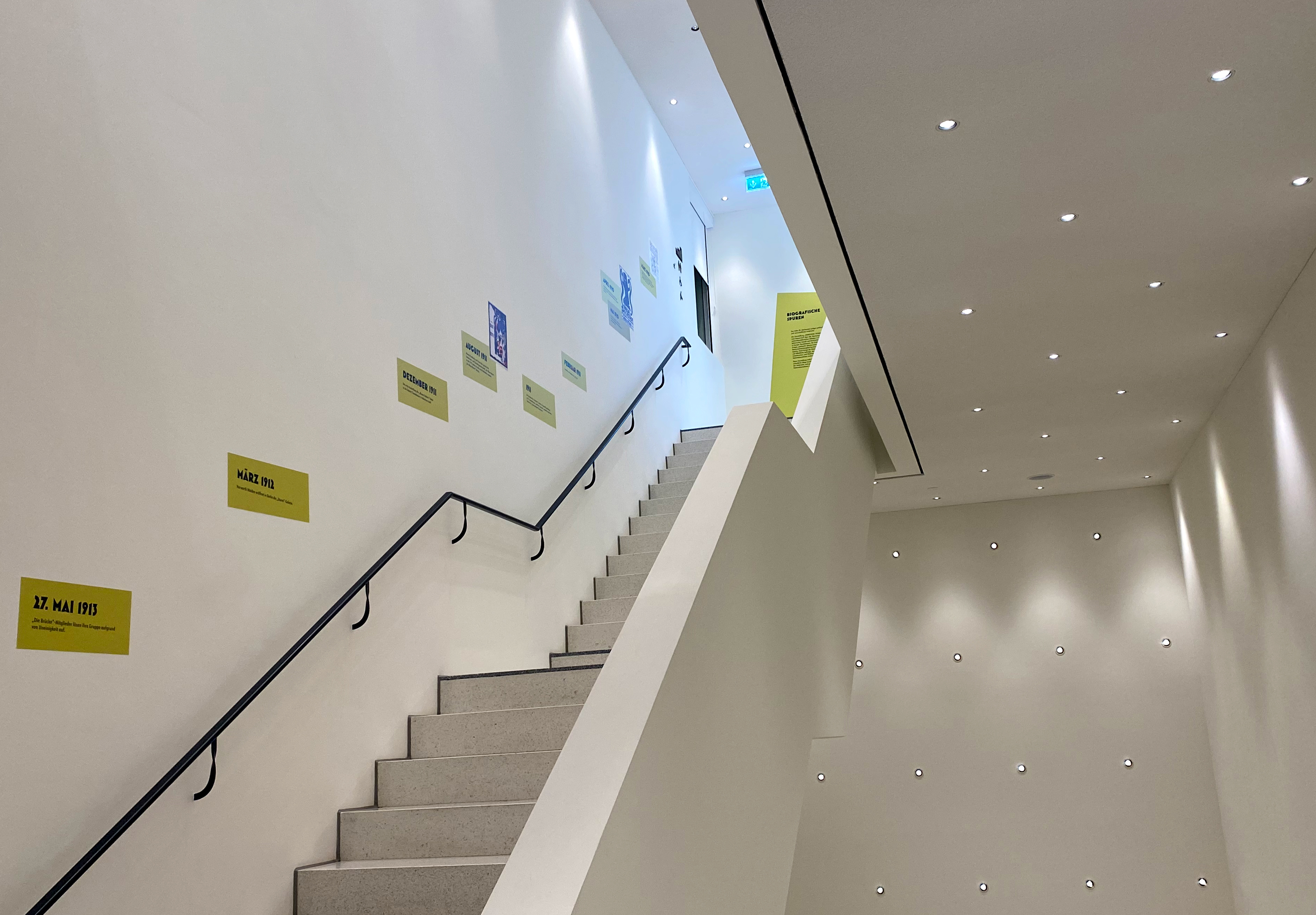 Weißes Treppenhaus. Gelbgrüne Flächen mit schwarzer Schrift bilden einen Zeitstrahl, den man über die Stufen ablaufen kann.