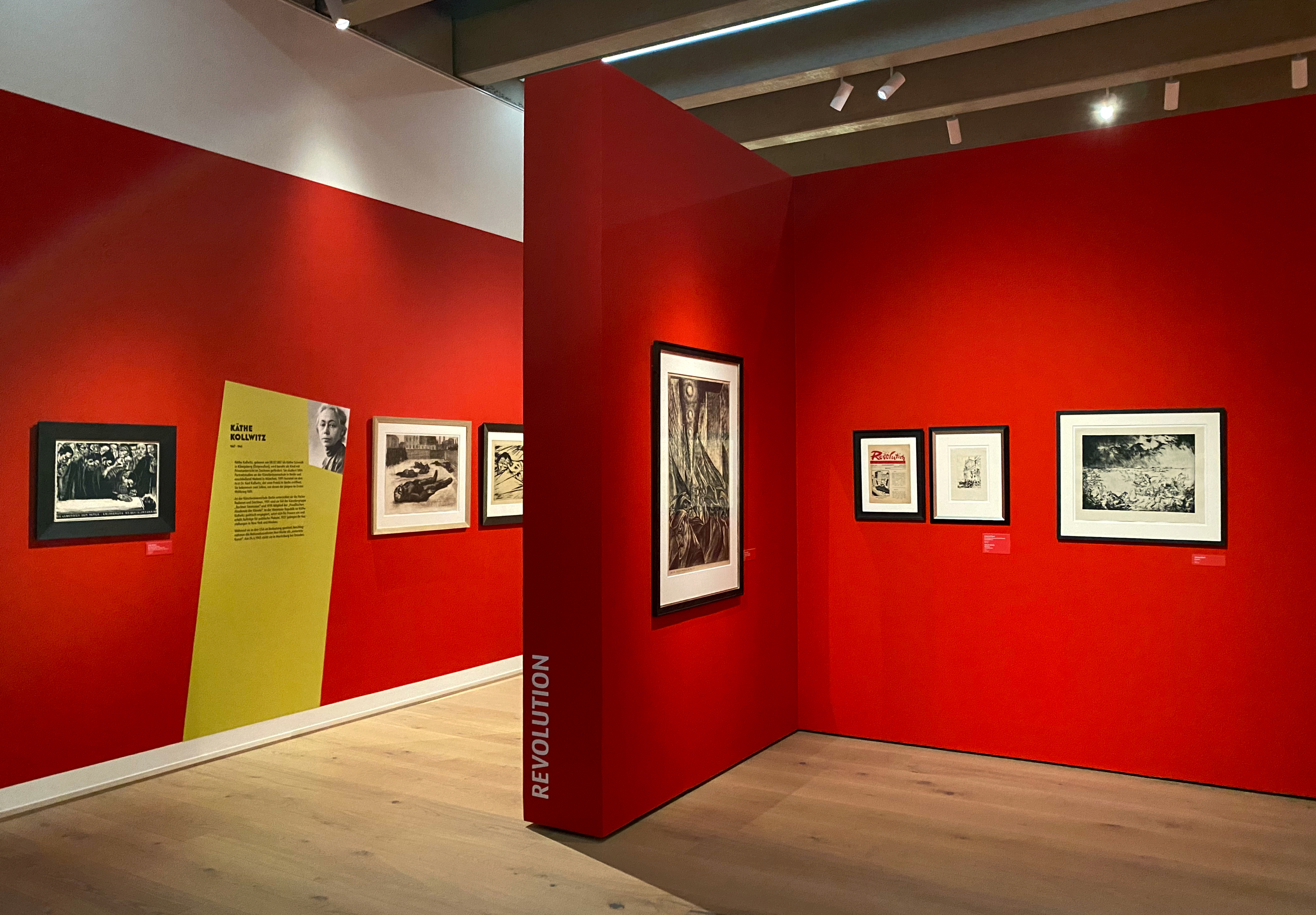 'REVOLUTION' steht gestürzt auf schmaler Seite von roter, verwinkelter Stellwänden. Bilder von unter anderem Käthe Kollwitz, hängen an den Wänden. Gelbgrüne Fläche, die vom Boden schräg nach rechts verläuft, beinhaltet Infotext zu Käthe Kollwitz, inklusive Foto.