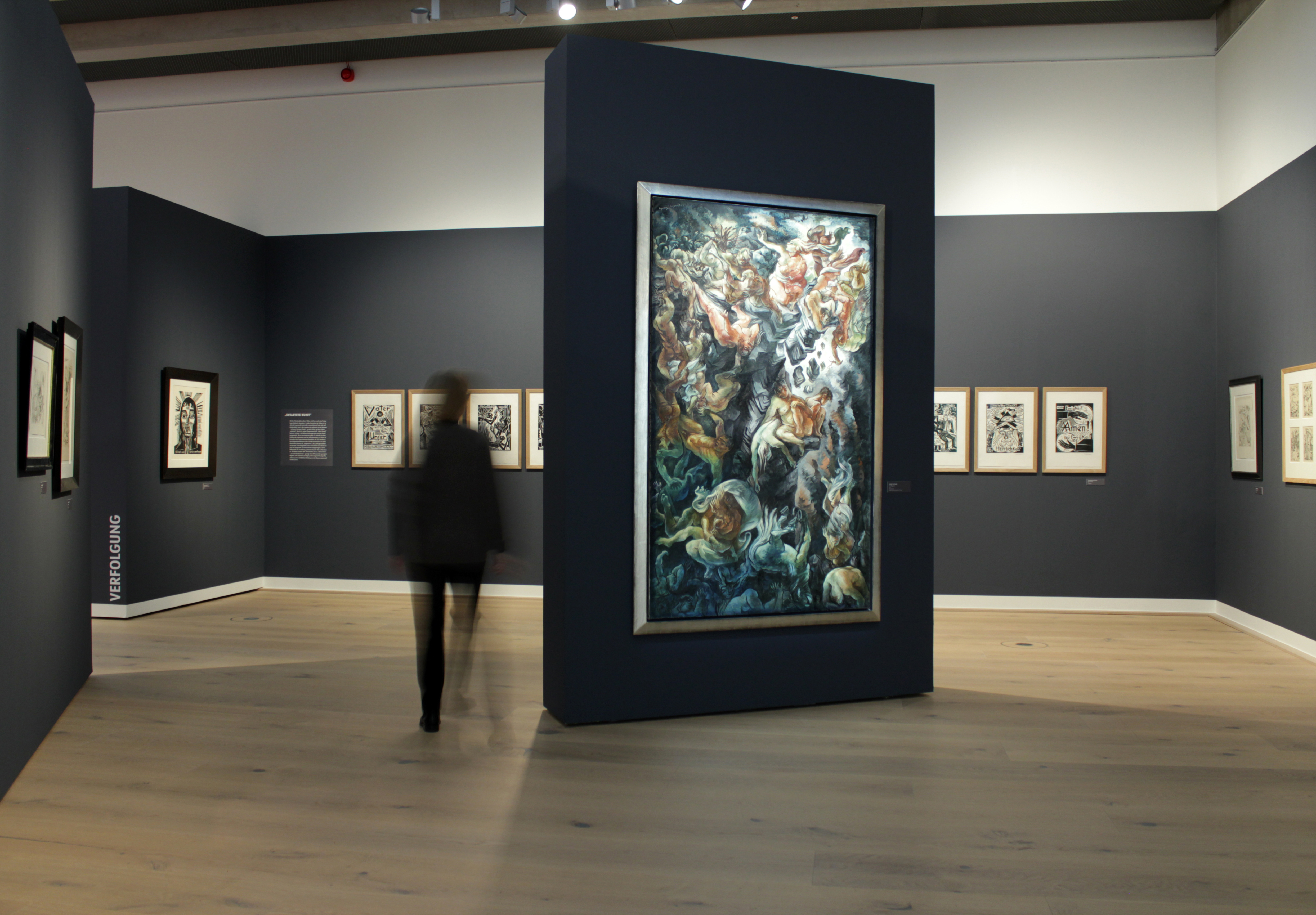 Dunkelgraublaue, freistehende Stellwand mit circa zwei Meter hohem und über einem Meter breitem Bild im Fokus. Motiv: 'Der Abgrund' von Georg Netzband von 1935. Umgeben von dunkelgraublauen Wänden mit weiteren Bildern. 'VERFOLGUNG' steht in weißen Versalien gestürzt auf der hinteren Wand.