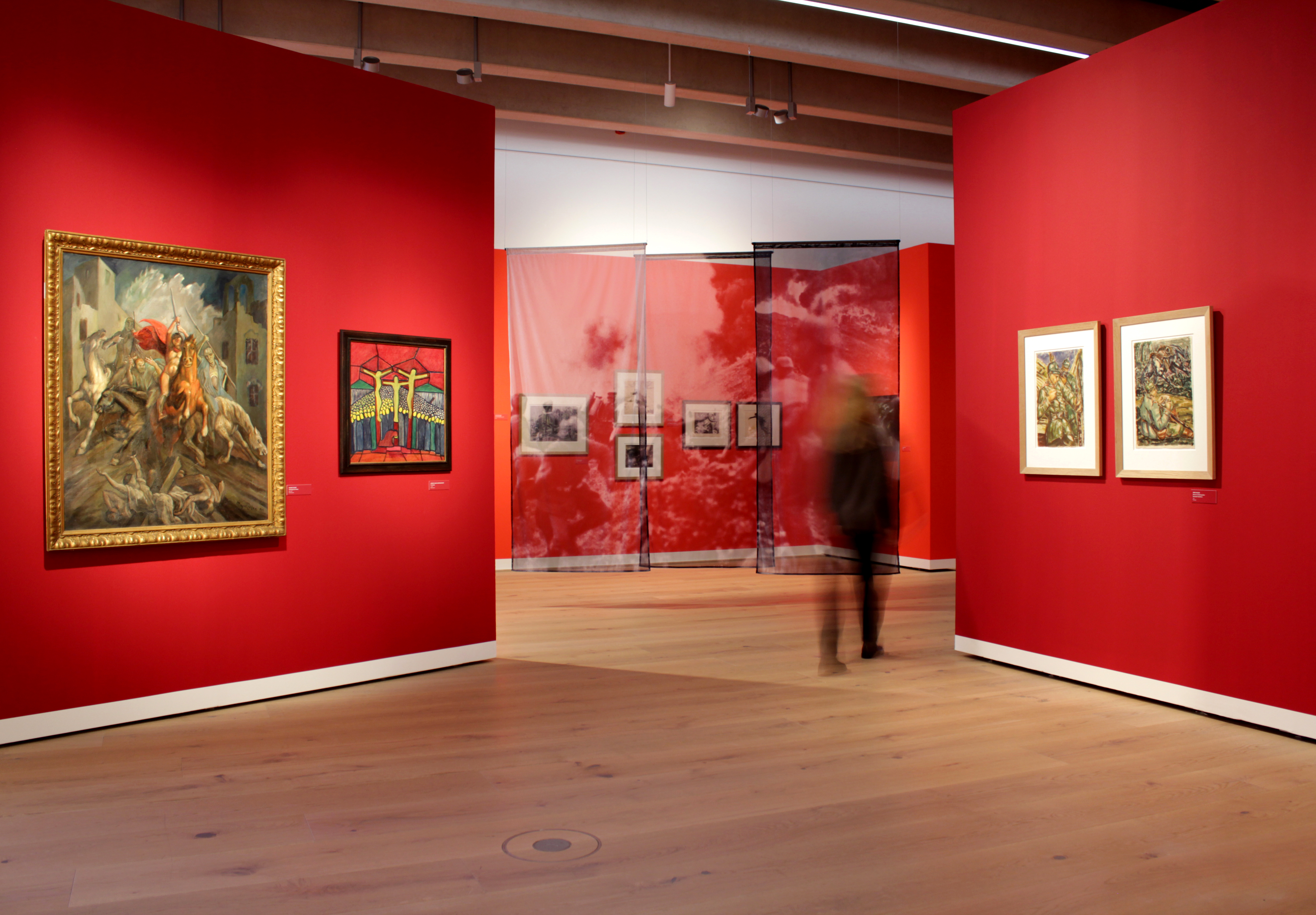 Zwei rote Wände, die aus dem Bildrand jeweils von links und rechts zur Mitte hin laufen. Pro Wand zwei gerahmte Bilder. Zentrum: Lücke zwischen beiden Wänden legt Blick auf drei schmale, hängende, bedruckte Textile frei. Dahinter sind 5 Bilder an Wand zu erahnen. Eine Person läuft durch die Ausstellung.