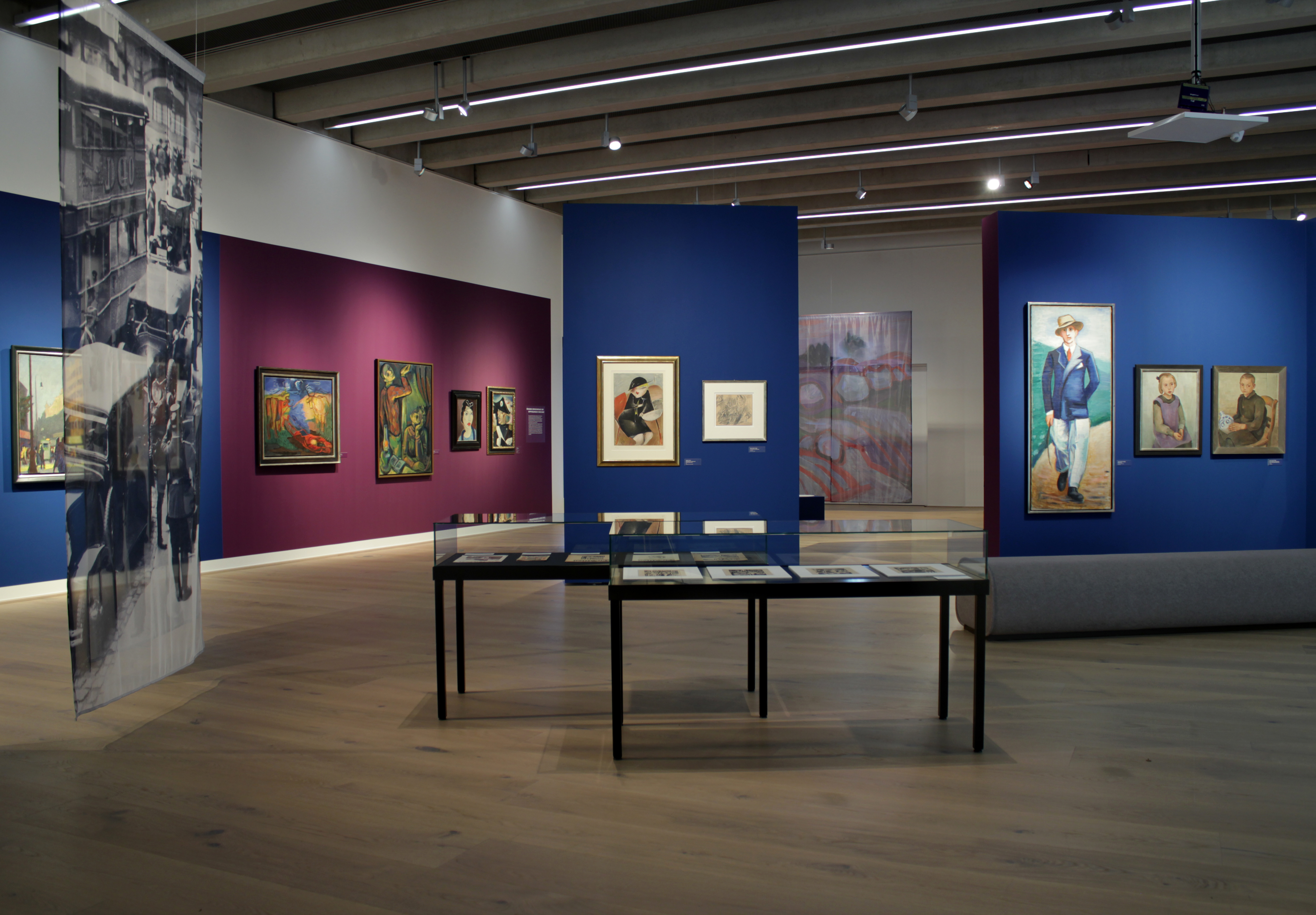 Zwei schwarze Tischvitrinen umgeben von blauen und lilanen Wänden mit Bildern. Im Hinter- und Vordergrund sind Teile von bedrucktem Gewebe zu sehen.