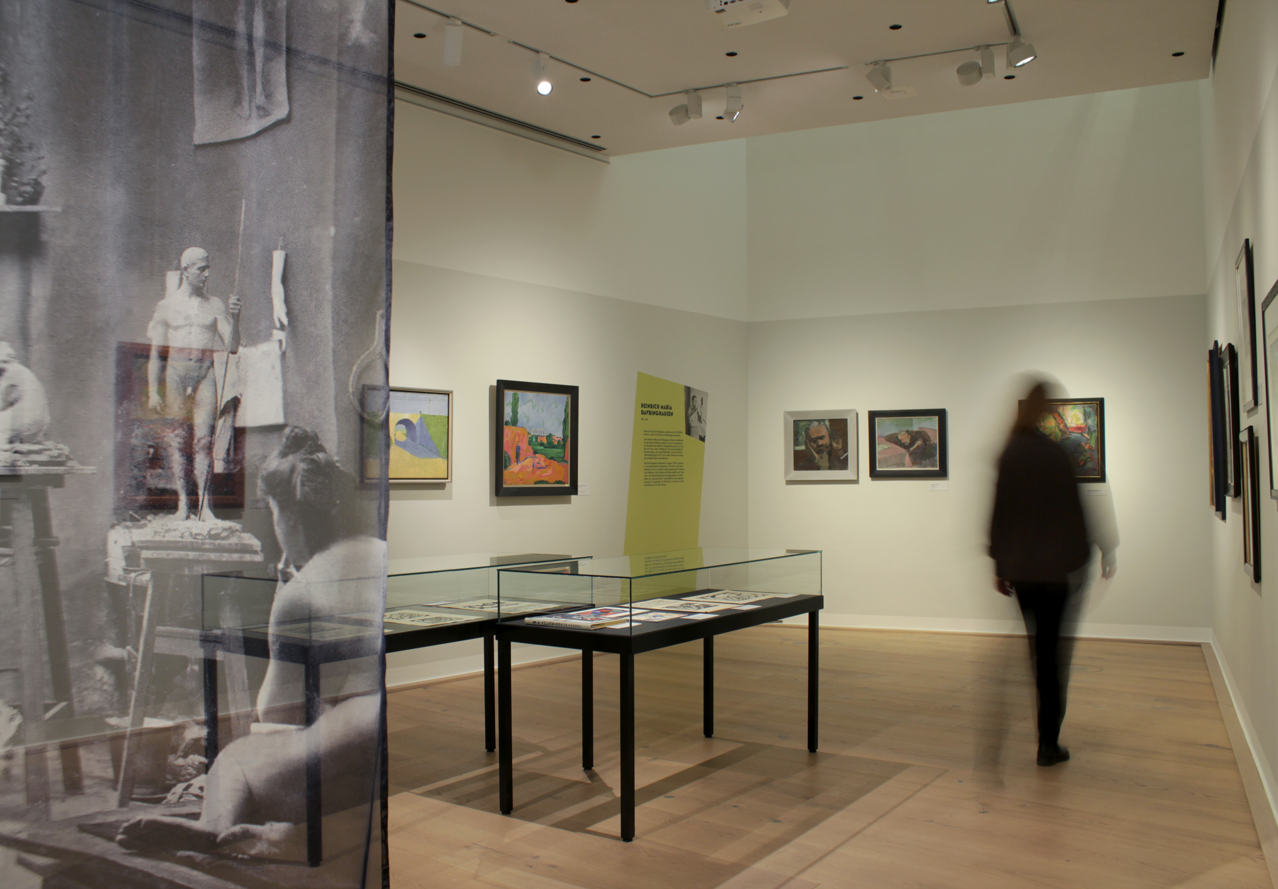 Zwei schwarze Tischvitrinen im Raumzentrum mit hellgrauen Wänden. Es hängen gerahmte Bilder. Hintere Ecke: Gelbgrüne Fläche mit dem Bereichstext. Blick in den Raum wird durch bedrucktes, transparentes Textil zu einem Drittel verdeckt. Eine Person läuft durch die Ausstellung.