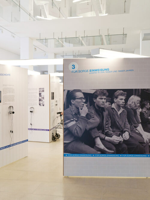 Ausstellungsraum mit weißen Ausstellungstafeln, auf denen Text und ein großes schwarz-weißes Foto von sitzenden Personen zu sehen sind