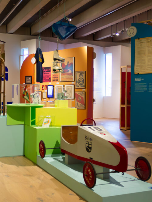 Blick in die Ausstellung mit einem roten und weißen Seifenkistenwagen auf einem Podest. Weitere Ausstellungsobjekte sind freistehend oder in Vitrinen auf farbigen Sockeln sowie von der Decke hängend platziert. An farbigen Ausstellungswänden hängen historische Wahlplakete.