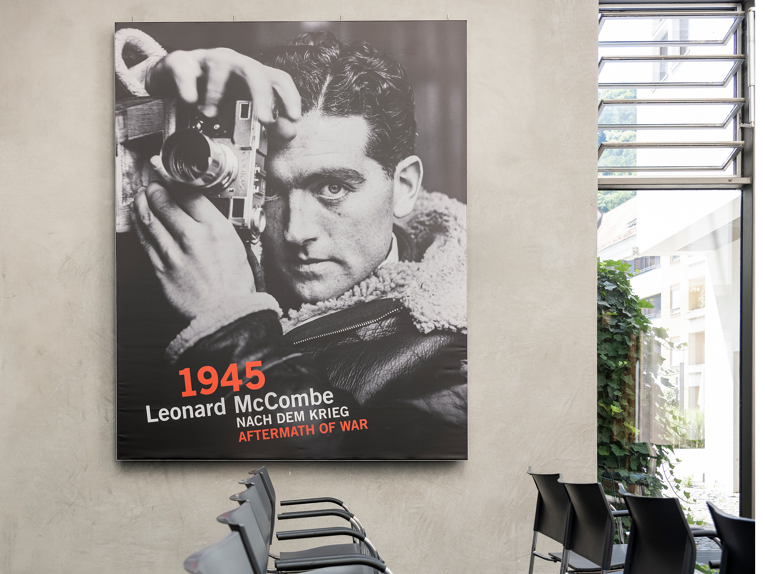 Schwarz-weiße Fotografie des Fotografen Leonard McCombe mit Kamera an einer Wand mit Text '1945 Leonard McCombe NACH DEM KRIEG', davor mehrere schwarze Stühle
