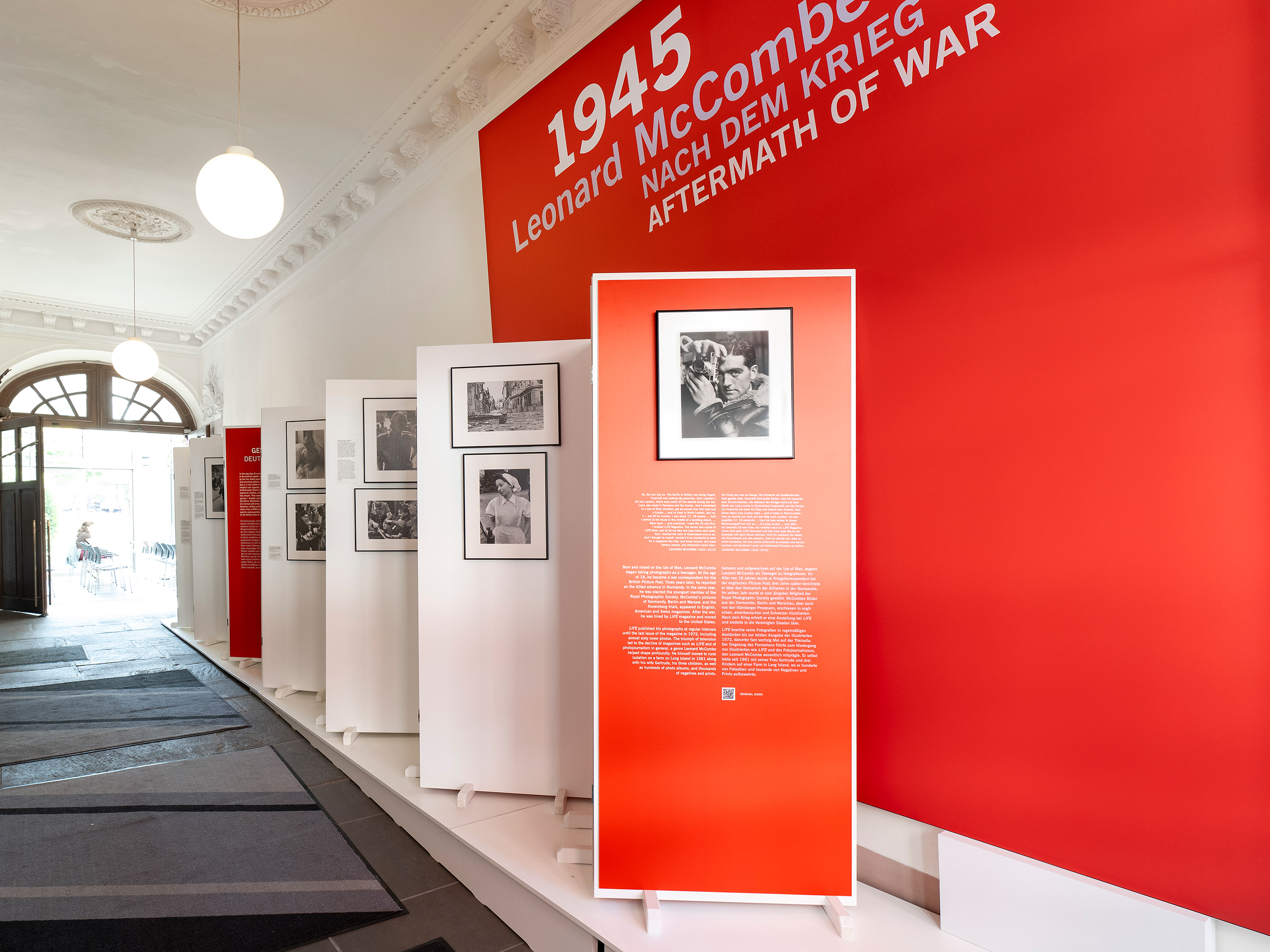 Ausstellung mit Fotografien und Texttafeln zu Leonard McCombe und dem Thema '1945 Nach dem Krieg' in einem hellen Raum mit roter Wand und hohen Decken