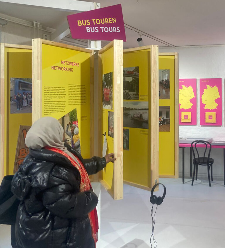 Person mit schwarzer Steppjacke und hellgrauem Kopftuch betrachtet gelbe Ausstellungstafeln mit Texten und Farbfotografien