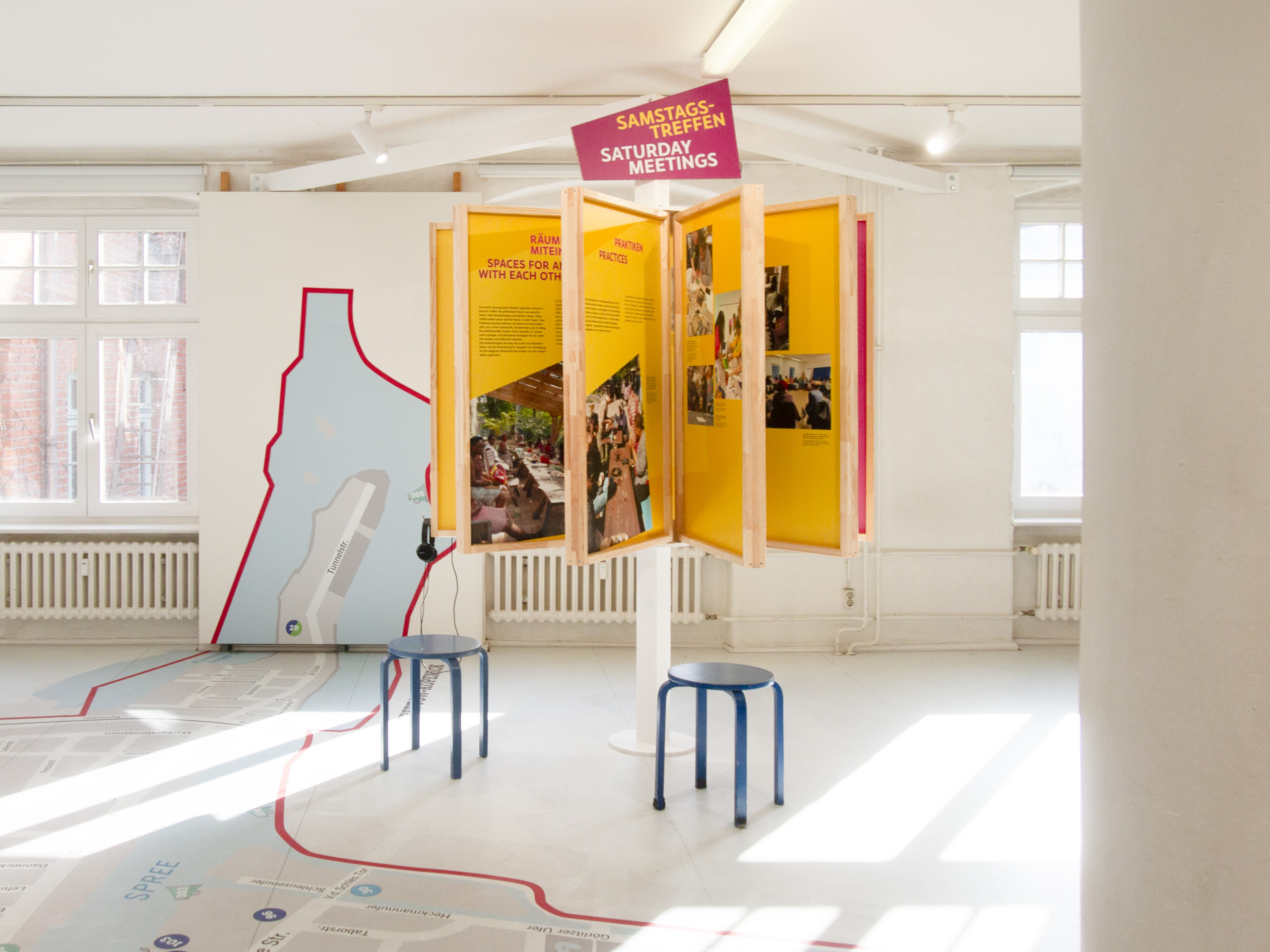 Ausstellung mit gelben Informationstafeln an einem drehbaren Ständer und zwei blauen Hockern in einem hellen Raum mit großen Fenstern und einem auf dem Boden aufgemalten Stadtplan. Themeninsel "Samstagstreffen"